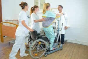 Medzinisches Personal übt mit Frau das Aufstehen aus dem Rollstuhl